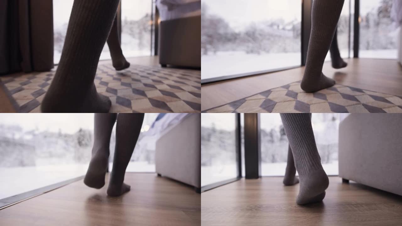一个特写镜头跟随一个穿着长统袜的女孩的脚，穿着袜子走过卧室里的一张床，卧室里有透明的墙壁窗户，后面是