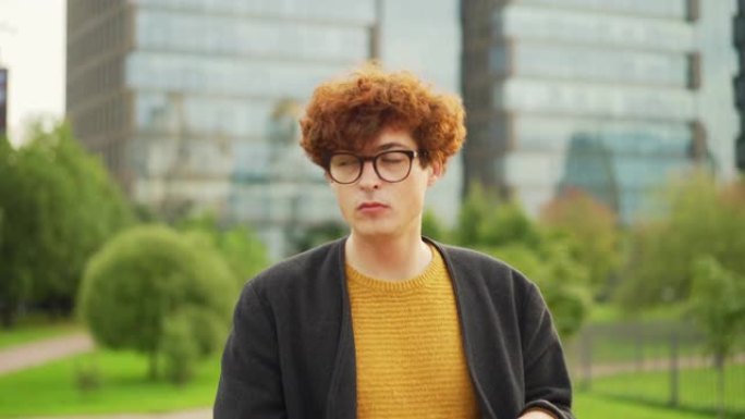 放大镜头，一个戴着眼镜、留着红色卷发的年轻人坐在户外的长椅上，在膝上放着公文包的笔记本电脑上打字，然