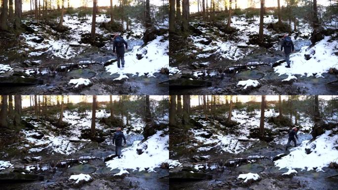 年轻的背包客在松树林的山河上走过石头。旅行时，无法识别的背包徒步旅行者在小溪上翻过岩石。寒假或假期的