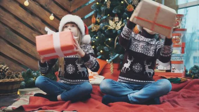 小双胞胎坐在圣诞树下，摇晃礼物盒子，聆听并找出里面的东西。