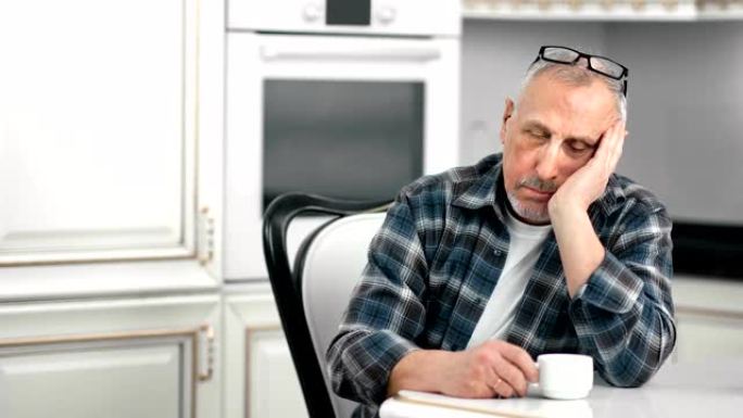 疲倦的衰老男性在白色厨房中等镜头喝茶时打瞌睡
