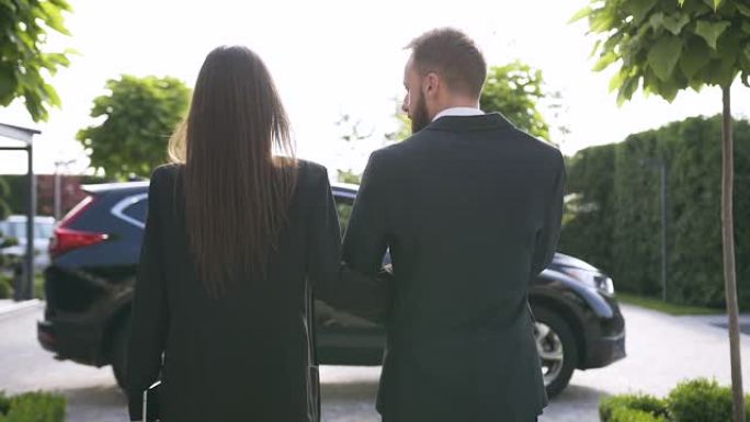 穿着正式服装走向黑色豪华车的成功的白人年轻夫妇的背景色