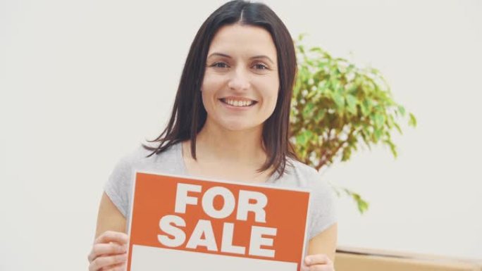 微笑的年轻美丽的女人拿着一个盘子，上面写着待售的单词，提供房屋出售并搬出。