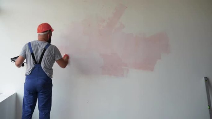 一名大胡子的男性建筑工人穿着工作服和防护头盔。用滚筒粉刷墙壁，总体平面图。