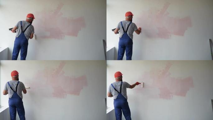 一名大胡子的男性建筑工人穿着工作服和防护头盔。用滚筒粉刷墙壁，总体平面图。