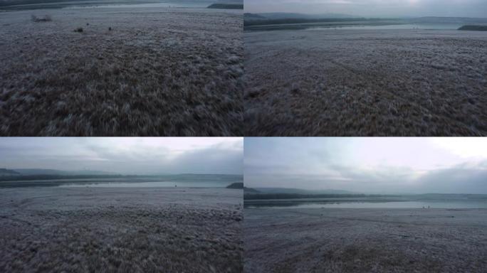 在英国南约克郡一个寒冷冰冷的冬季早晨日出期间，航拍画面飞越一个仍然霜冻的湖泊和冰冻的草地