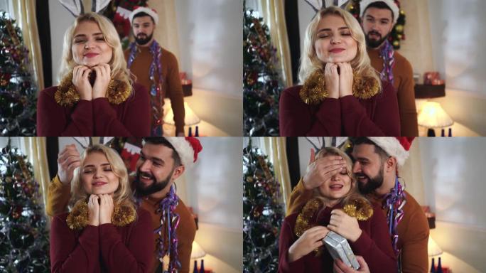 戴着圣诞帽的大胡子高加索人从背景中来到漂亮的金发女人，闭上眼睛，赠送礼品盒。快乐的丈夫为可爱的妻子做