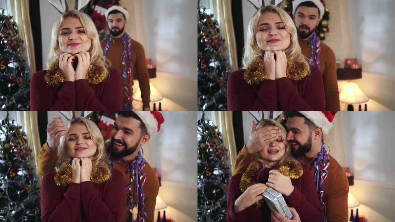 戴着圣诞帽的大胡子高加索人从背景中来到漂亮的金发女人，闭上眼睛，赠送礼品盒。快乐的丈夫为可爱的妻子做