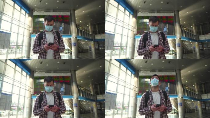 戴着口罩的男子游客在机场航站楼使用智能手机从抵达离境板检查航班。旅行时冠状病毒 (新型冠状病毒肺炎)