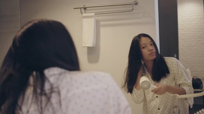 亚洲美女在酒店内部背景的浴室里用吹风机在镜子上梳理和造型头发。4k慢动作。
