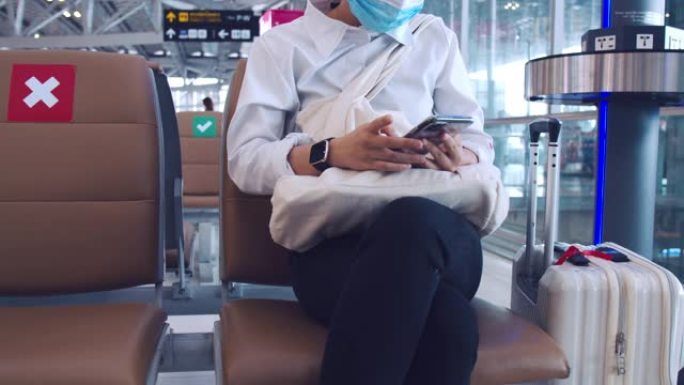 女商人戴着外科口罩保护，并在机场候机楼坐在椅子上使用智能手机检查社交媒体供稿，并在新型冠状病毒肺炎大