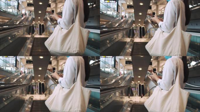 亚洲女商人戴着外科口罩保护，在机场航站楼的自动扶梯上使用智能手机打字短信，并在新型冠状病毒肺炎大流行