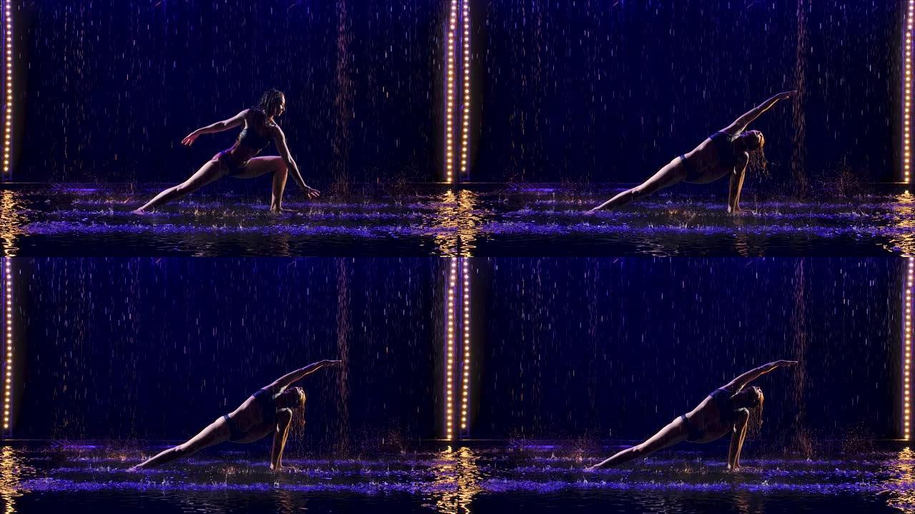 年轻女子在工作室蓝光和水雨下进行瑜伽练习。水滴中的女性身体。在黑色背景的工作室里拍摄的照片。慢动作