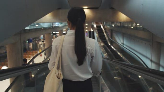 年轻女性戴着外科口罩保护，在机场航站楼的自动扶梯上使用智能手机，并在新型冠状病毒肺炎大流行期间保持社