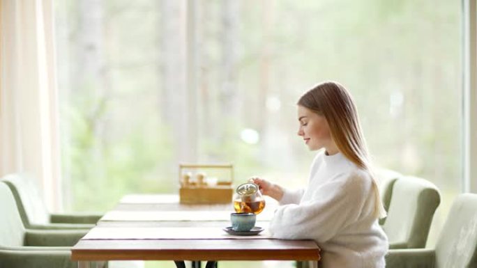 侧视图缩小镜头，美丽的年轻女子独自坐在咖啡桌，喝着茶，幻想着她的脸上的微笑