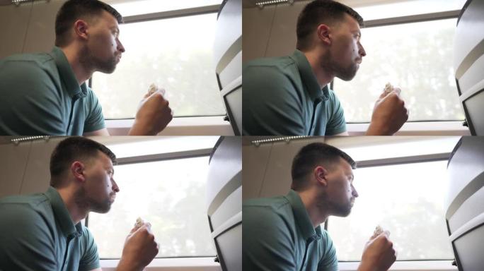 饥饿的家伙正在坐在窗边的火车车厢里吃快餐。旅行铁路。不良的不健康习惯。垃圾食品走了。一个人乘火车旅行