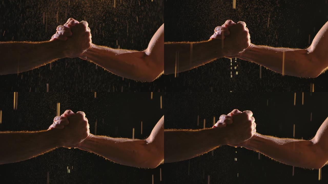 两个肌肉发达的男人在雨滴和许多飞溅的黑色背景上握手。慢动作。特写