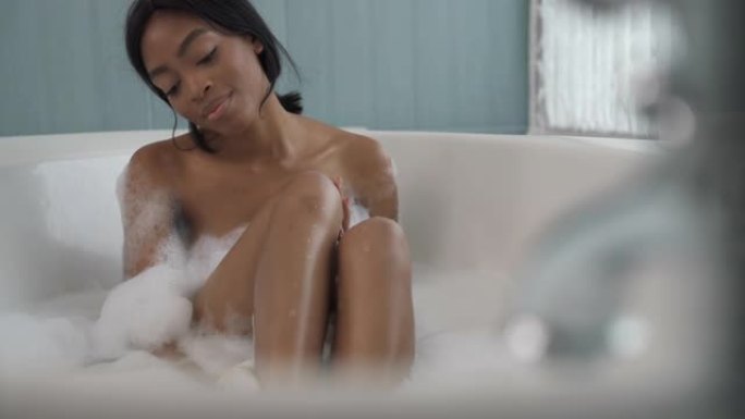 非裔美国妇女坐在泡泡浴中，用去角质手套洗自己。水疗和护肤概念。左平底锅射击