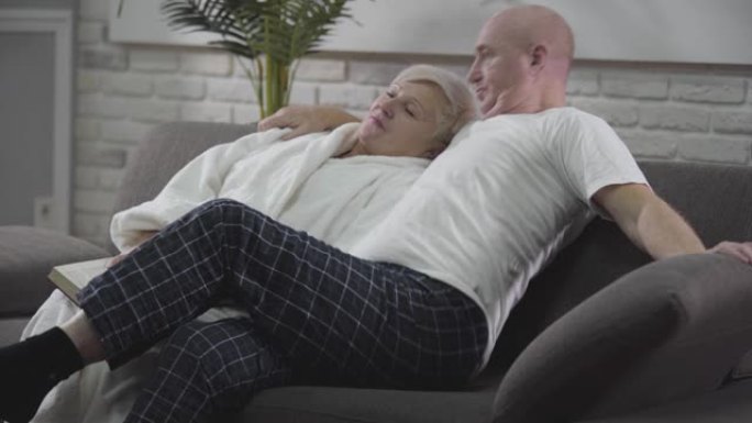 高个子高加索秃头男子坐在沙发上，拥抱妻子，说话和打手势的侧视图。恋爱中的成熟夫妇一起度过安静的夜晚。