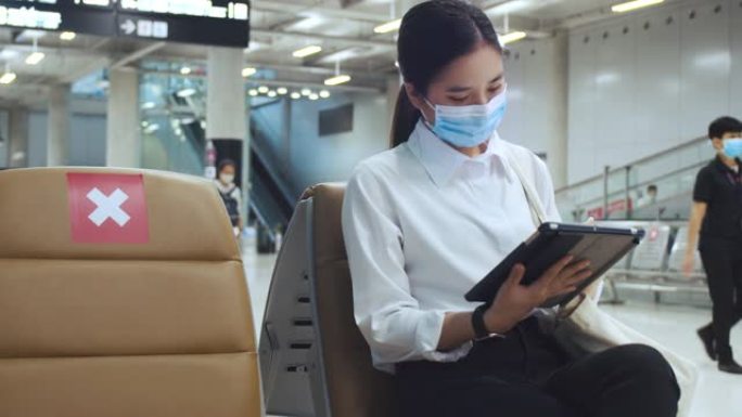 戴着外科口罩的女人面部保护，坐在机场航站楼的椅子上使用平板电脑检查社交媒体，并在新型冠状病毒肺炎大流
