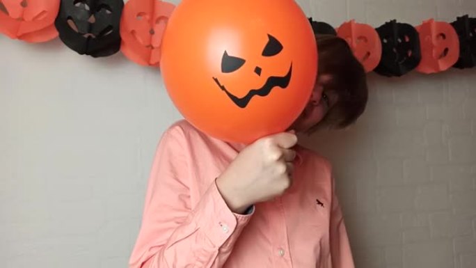 一个男孩把脸藏在橙色南瓜气球后面，家里有Jack'O灯笼的脸，玩耍，再次露面