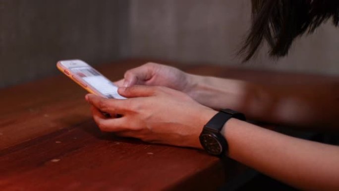 4k亚洲女性手使用白色智能手机，向下滚动手机，戴着黑色手表，手放在木桌上，在家，晚上，社交媒体，手机