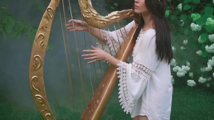 漂亮的精灵女孩玩金色老竖琴。春天开花花园里的公主。