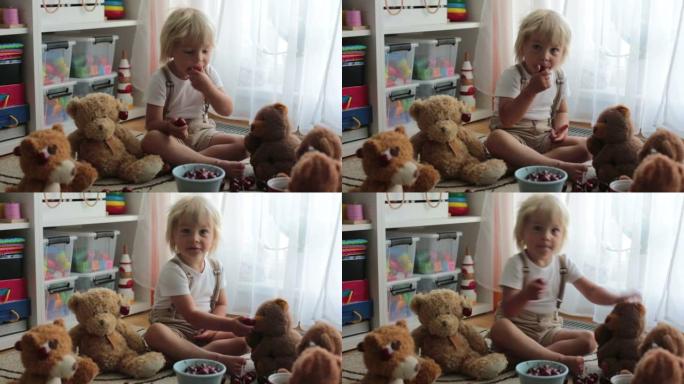 蹒跚学步的金发孩子，可爱的男孩，在家用泰迪熊吃樱桃
