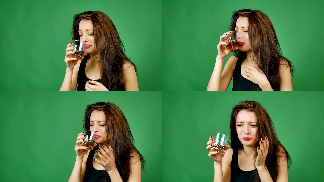 年轻的黑发悲伤女孩，留着长长的黑发，正在工作室里从绿色背景的玻璃杯里喝威士忌，女人的酗酒，酗酒的概念