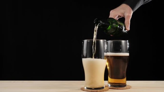 新鲜啤酒。手将美味的精酿啤酒倒入第二杯啤酒杯垫中，从棕色瓶子倒入黑色背景下的木桌中。带有水滴，气泡和