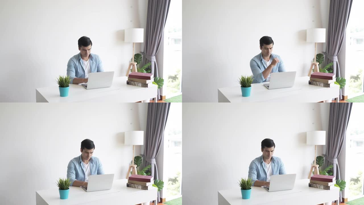 穿着蓝色衬衫的快乐休闲男人坐在桌子上，在客厅用笔记本工作