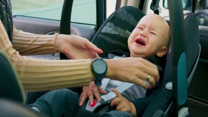 特写哭泣的男婴坐在车内的婴儿汽车座椅上。妈妈系好安全带，抬起把手，关上门。婴儿透过玻璃看着微笑。慢动