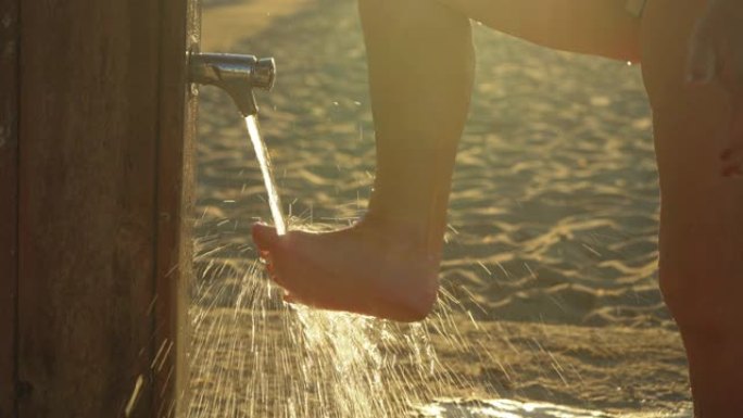 年长的女人在海滩淋浴下用沙子洗脚，喷水细节，早晨的太阳背景