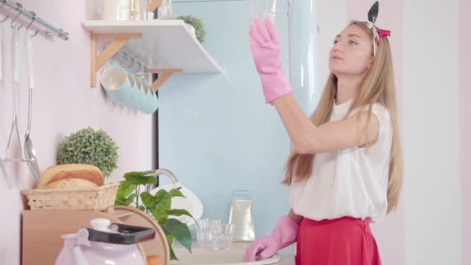 疲惫的年轻家庭主妇在厨房洗碗，欣赏干净的水杯。穿着粉红色家政手套的美丽白人妇女的肖像在家打扫卫生