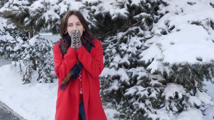 美丽的年轻黑发女人，穿着红色外套，围巾，连指手套，微笑着冻结试图热身，冬天下雪的日子在外面吹手