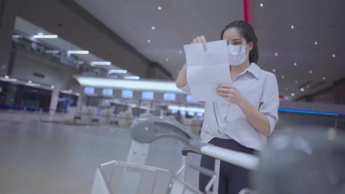 亚洲年轻女性戴着口罩检查机票旅行路线预订文件纸，在空荡荡的机场航站楼，女士独自旅行，前台有行李手推车