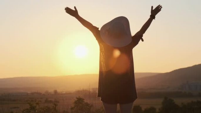 穿着衣服和帽子的幸福女人的剪影站在美丽的风景田野上，背景是明亮的日落和欣赏自然的慢动作，举手晒太阳。