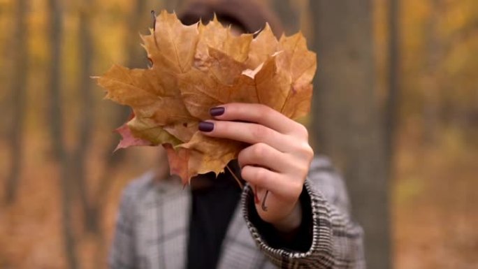 一位戴着眼镜的年轻女子躲在秋天森林中的一棵大叶树后面。女孩对着镜头伸出树叶。慢动作