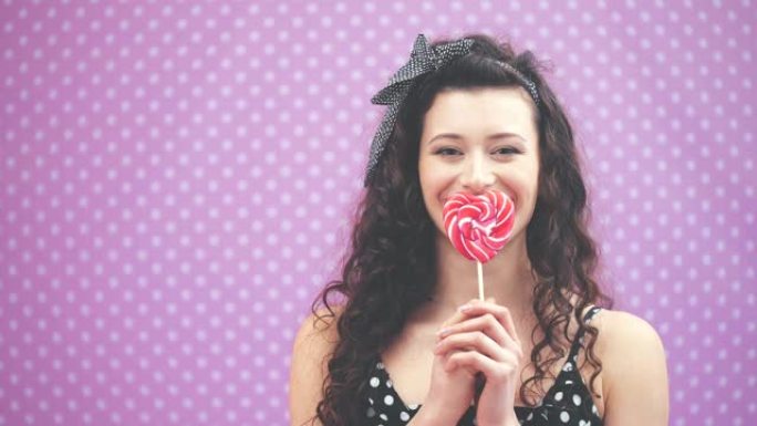 可爱的年轻卷发女孩站着，嘴前的棍子上拿着甜美的红色棒棒糖。