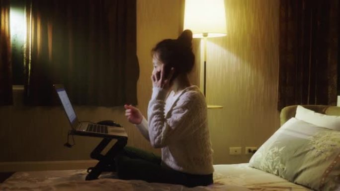 一个女人在卧室里用笔记本和手机上班，晚上开灯。