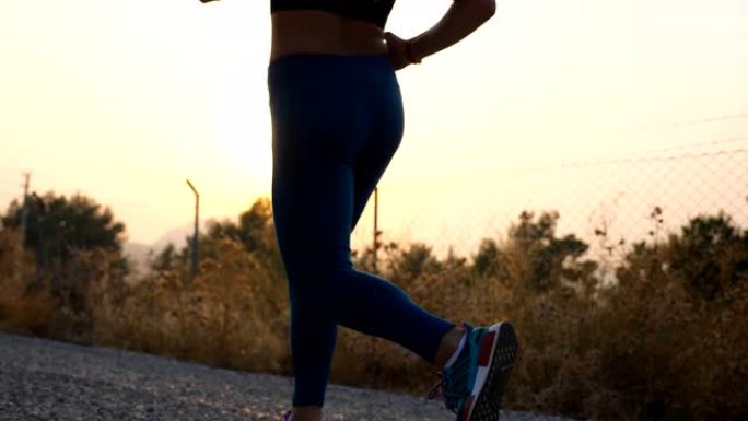 年轻健康女孩的脚在乡村道路上慢跑，背景是日出。室外跑步的苗条女性双腿。运动的女人在清晨锻炼。积极生活