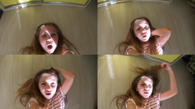 可爱的小女孩的女性脸，长长的金发躺在房间的地板上，在GoPro上拍摄自己。快乐的孩子在家里玩得开心并