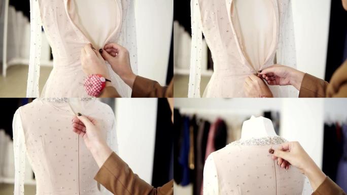 在明亮的工作室，无法辨认的女裁缝将粉底服装系在闪闪发光的婚纱上。女人的双手合拢，将衣服的后背拉上拉链