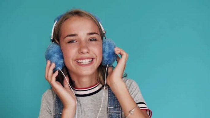 可爱的微笑女孩戴着蓝色大耳机在工作室里听音乐。把手放在耳机上。可爱的少女通过蓝色背景上的耳机听音乐