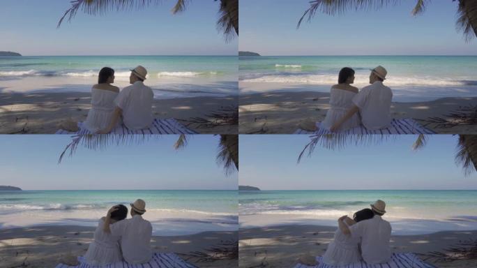 恋爱中的年轻亚洲夫妇坐在热带海滩的棕榈树下，在度蜜月的时候看着美丽的夏日大海。暑假和假期概念。