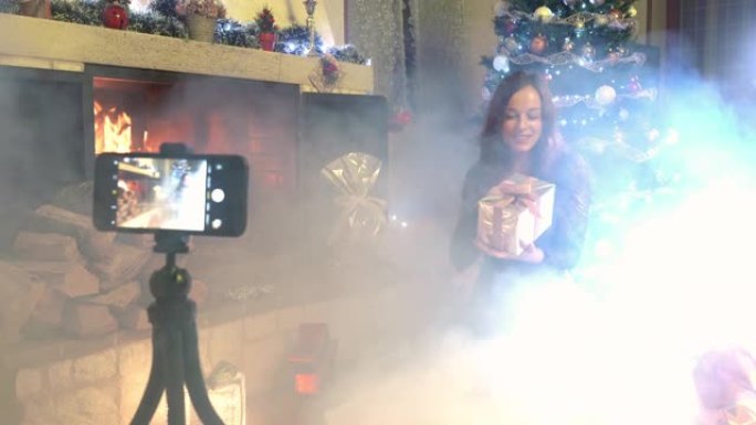 燃烧的壁炉和发光的圣诞树令人惊叹的圣诞气氛，漂亮女人为她的个人博客录制赠送视频，为带有粉色丝带的彩票