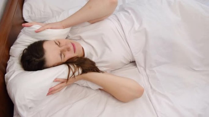 年轻女子躺在白色床上，用枕头遮住头，因为太大的烦人噪音使她无法入睡。被激怒的女孩因大声噪音问题而不安