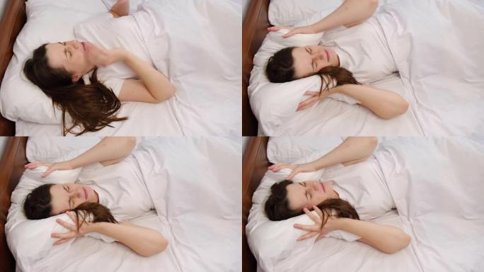 年轻女子躺在白色床上，用枕头遮住头，因为太大的烦人噪音使她无法入睡。被激怒的女孩因大声噪音问题而不安