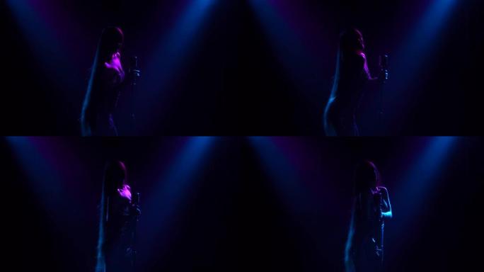 一位专业歌手在黑暗中用蓝灯在舞台上唱一首歌。一位穿着闪亮连衣裙的年轻女子在一场真正的音乐会上对着老式