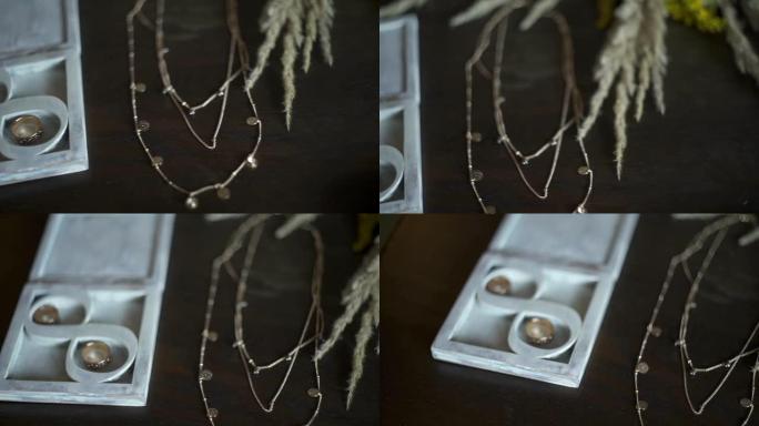 深色木制桌子上的新娘配饰: 新娘和新郎的戒指，木盒中的珠宝，吊坠，金链，花束。特写慢动作。婚礼当天。